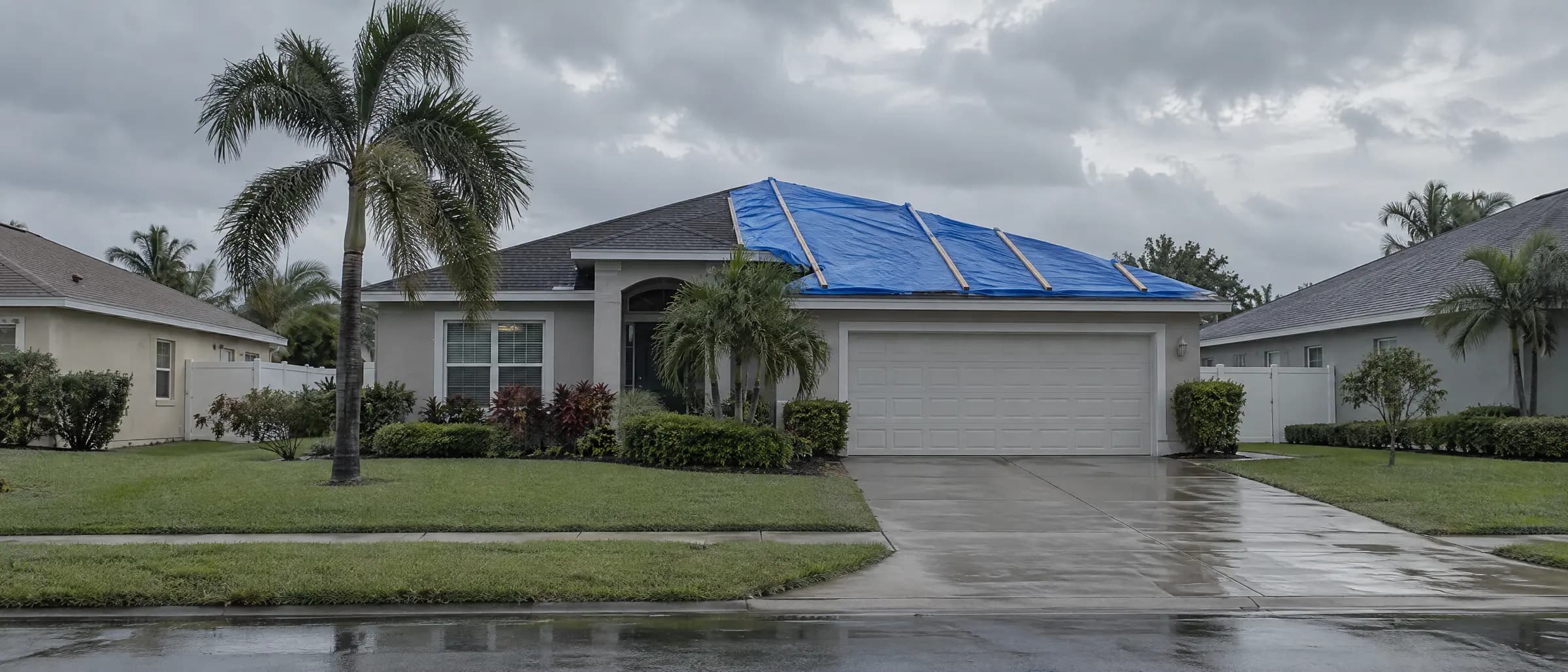 A blue tarp strapped over a storm-damaged roof, seen from the street on an overcast morning